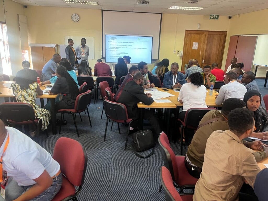 Several groups of participants seated classroom style, at the 2024 AMR Symposium, South Africa. A lead presenter is standing at the front of the room, next to a projector. 