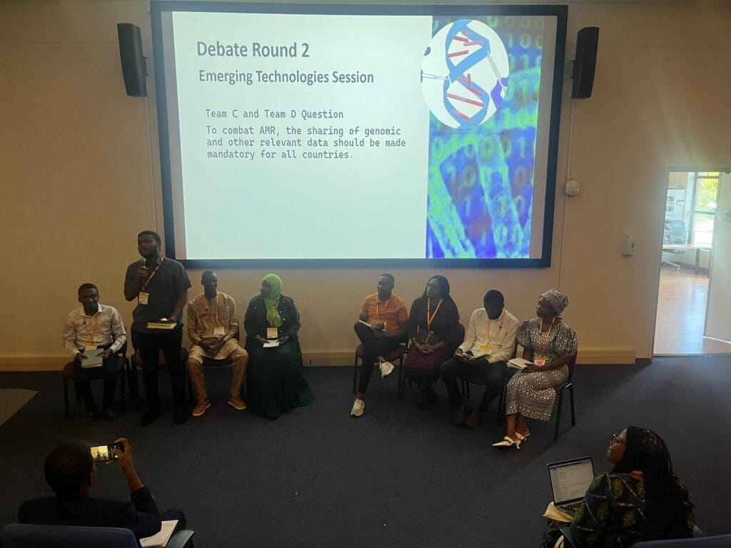 Seven panelists seated in a row underneath a screen. The screen is projecting a slide on the topic of emerging technologies in AMR. The panelists are facing a group of delegates. One speaker is standing and holding a microphone. 