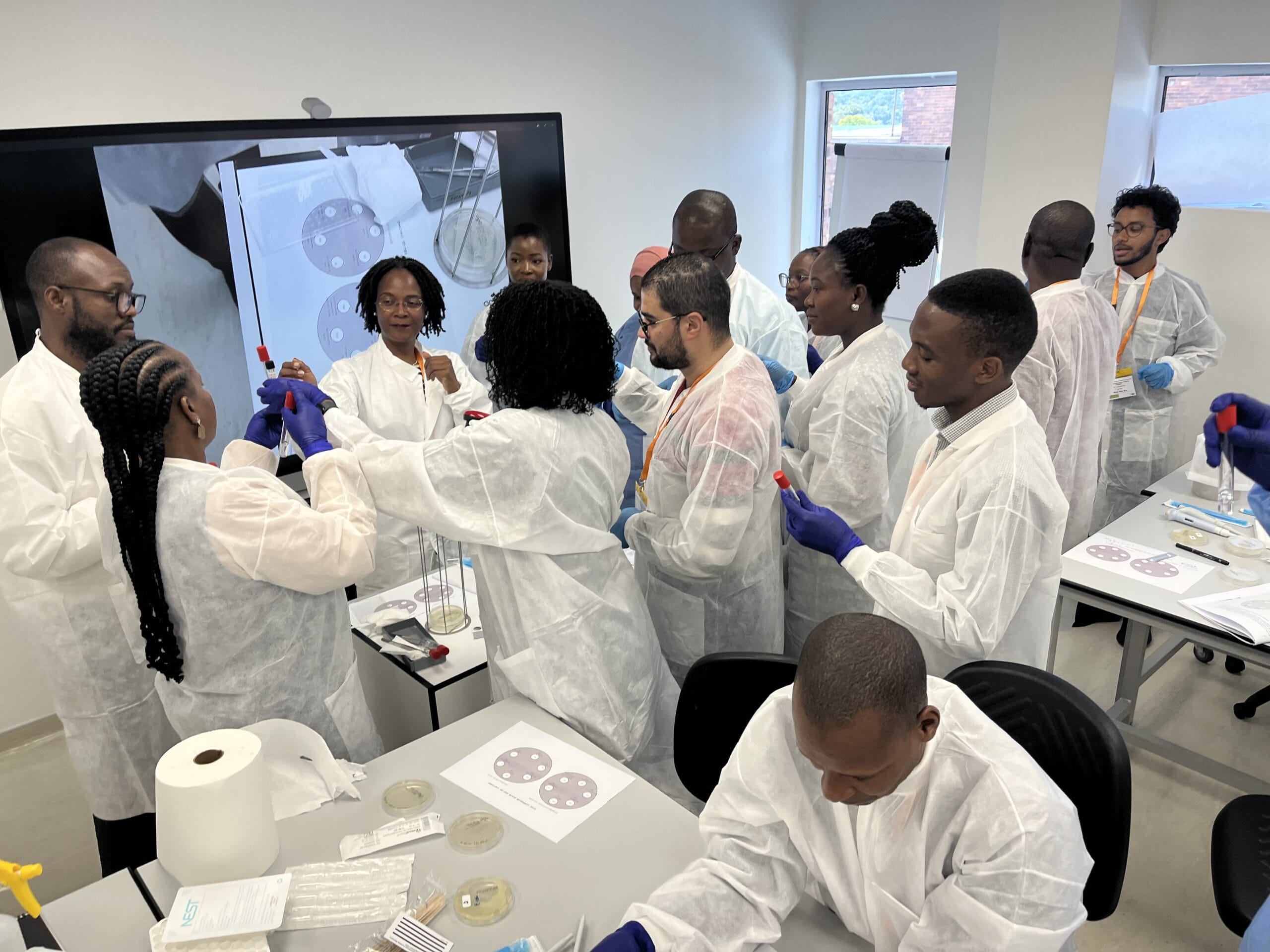 A group of course participants in training laboratory wearing white lab coats looking at, and comparing, test tubes. 