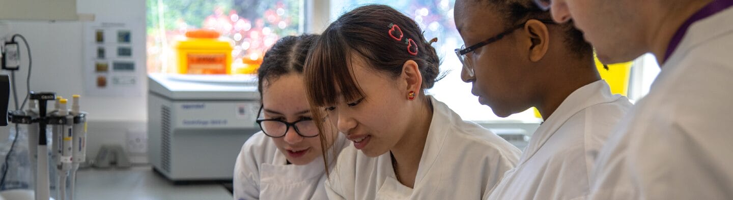 A group of young people are looking at something in a lab, they are wearing lab coats.